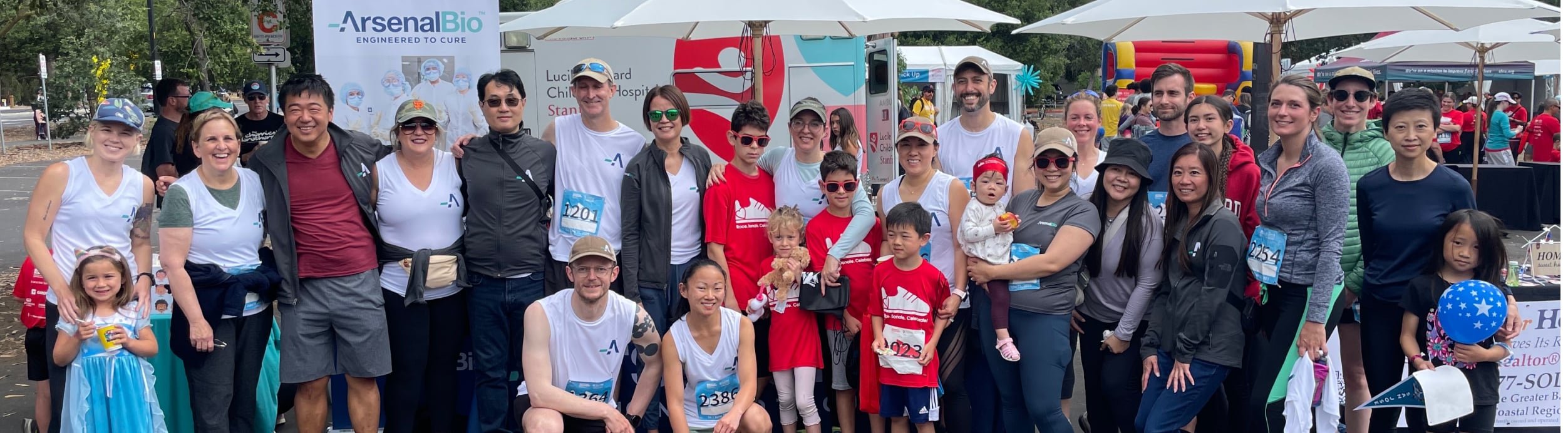 about 25 people, adults and children, at an outdoor event, many with running bibs and shirts with Arsenal's "A" logo on them.